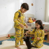 Two children wearing matching yellow dinosaur-print pajamas play with a small black kitten in a brightly lit children's bedroom.